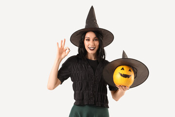 Beautiful young happy woman dressed as witch for Halloween with pumpkin showing OK gesture on white background