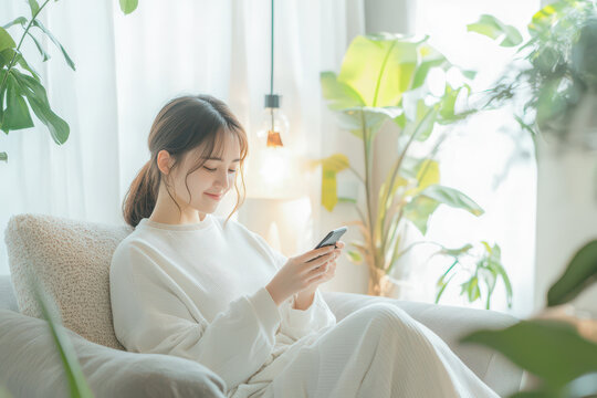 Young asian woman in cozy attire sits comfortably on a sofa, using her smartphone in a bright, plant-filled living room.
