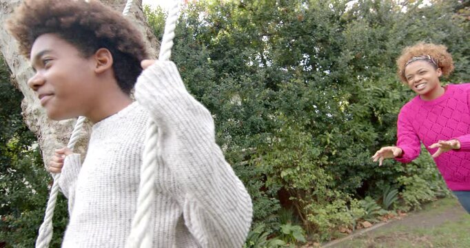 Happy african american mother pushing her son on tree swing in garden, slow motion