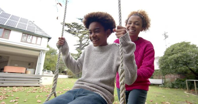 Happy african american mother pushing her son on tree swing in garden, slow motion