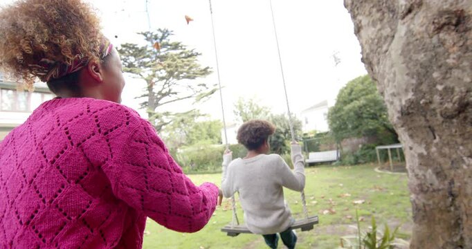 Happy african american mother pushing her son on tree swing in garden, slow motion