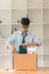 A young man in a blue shirt and tie unpacks a box at a wooden table, focused on organizing items in a minimalist room with shelves.