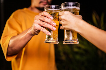 A man's hand and a woman's hand hold beer glasses in a cozy, lively evening party setting. Family and friends gather under the night sky, sharing joyful moments and laughter.