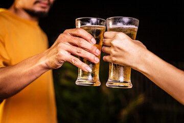 A man's hand and a woman's hand hold beer glasses in a cozy, lively evening party setting. Family and friends gather under the night sky, sharing joyful moments and laughter.