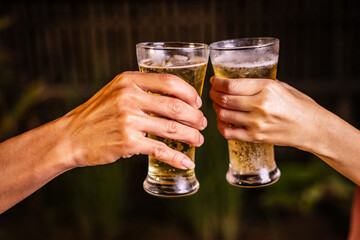 A man's hand and a woman's hand hold beer glasses in a cozy, lively evening party setting. Family and friends gather under the night sky, sharing joyful moments and laughter.