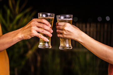 A man's hand and a woman's hand hold beer glasses in a cozy, lively evening party setting. Family and friends gather under the night sky, sharing joyful moments and laughter.