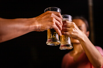 A man's hand and a woman's hand hold beer glasses in a cozy, lively evening party setting. Family and friends gather under the night sky, sharing joyful moments and laughter.