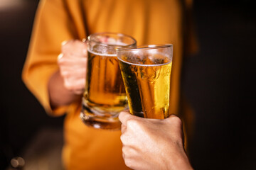 A man's hand and a woman's hand hold beer glasses in a cozy, lively evening party setting. Family and friends gather under the night sky, sharing joyful moments and laughter.