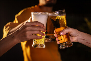 A man's hand and a woman's hand hold beer glasses in a cozy, lively evening party setting. Family and friends gather under the night sky, sharing joyful moments and laughter.