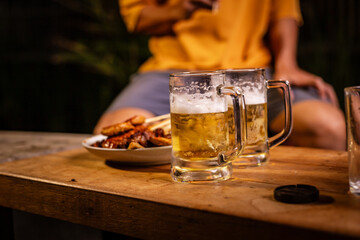 A man's hand and a woman's hand hold beer glasses in a cozy, lively evening party setting. Family and friends gather under the night sky, sharing joyful moments and laughter.