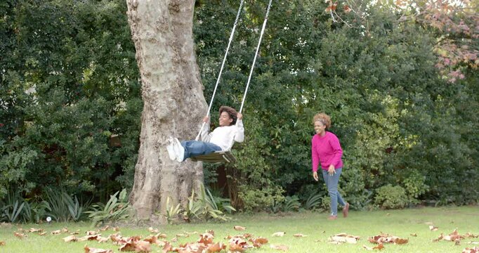 Happy african american mother pushing her son on tree swing in garden, in slow motion