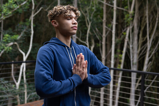 Young man meditating on balcony in peaceful forest setting, wearing blue hoodie
