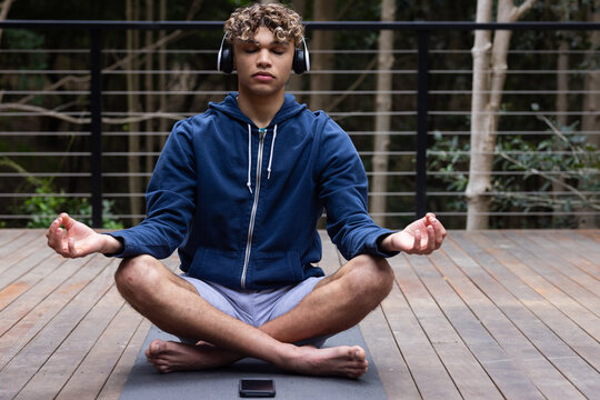 Young man meditating on balcony on yoga mat, wearing headphones, eyes closed