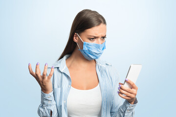 Discontented Woman Using Cellphone Reading News About Coronavirus Standing On White Studio Background, Wearing Medical Mask