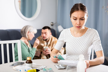 Fototapeta premium Portrait of dissatisfied young woman sitting at table choosing medicines for her sick husband, while her mother-in-law giving drugs to him