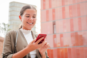 Copy space. Smiling young woman using a cellphone outdoors, checking messages or social media in an urban setting. Female holding a phone in casual business attire with a modern building background