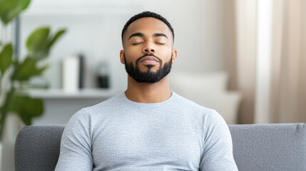 Man with beard sits peacefully on gray sofa, eyes closed, in bright living room with plants and books in background