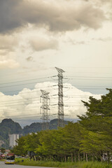 Tall power transmission tower with multiple cables stretch across the sky, set against a cloudy backdrop. Lush greenery and trees fill the foreground, contrasting with the industrial structures above.