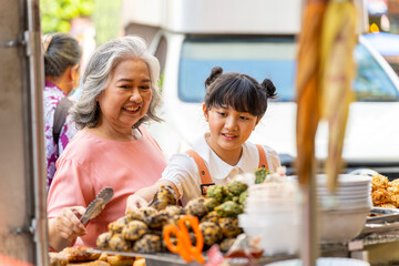 Happy Asian family grandmother and grandchild girl enjoy and fun outdoor lifestyle shopping and eating street food together during travel city street market Chinatown on summer holiday vacation.
