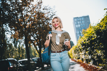 Photo of positive young blonde girl hold gadget wear khaki outfit tied shoulders sweater walk park...