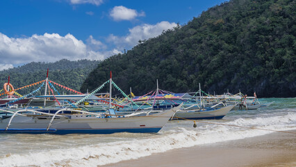 Traditional Filipino bangka boats are moored near the ocean shore. The masts are decorated with multicolored flags. Colorful awnings above the deck.The waves are foaming on the sandy beach.Green hills