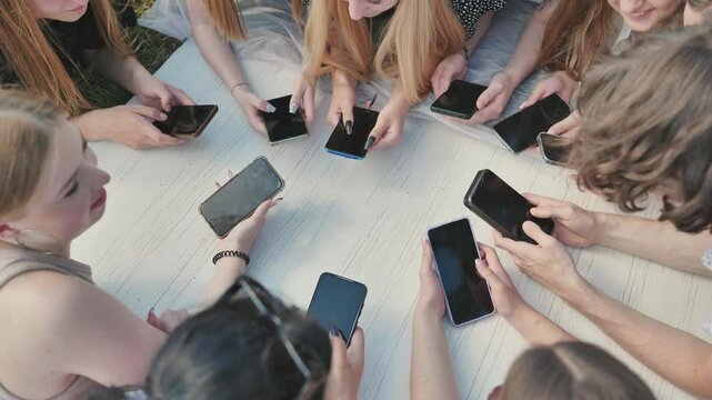 Group of teenagers lying on grass using smartphones together around white table.