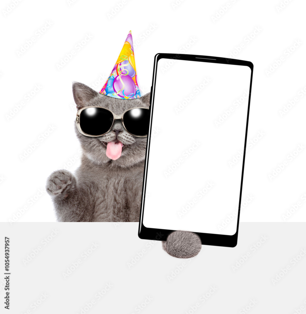 Poster Happy kitten with tongue out wearing party cap shows big smartphone with blank screen above empty white banner. Empty space for text. isolated on white background