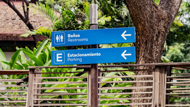 Blue bilingual signs pointing to restrooms and parking in Spanish and English, set against a lush outdoor background, ideal for tourism and wayfinding themes.