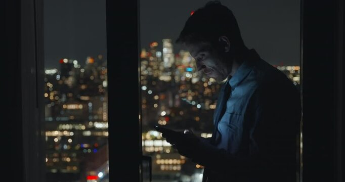 Young tired and angry businessman typing on his mobile phone in anxious manner. Annoyed or discontent about work or project. Standing at hotel office or apartment window overlooking the city at night