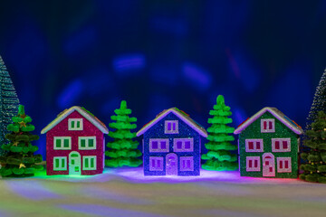 On a dark blue background among green fir trees there are multi-colored houses with illumination