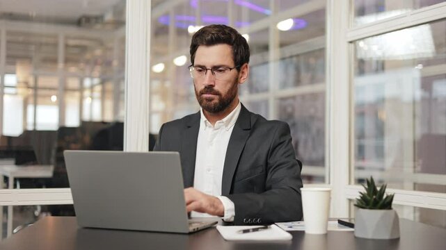 Satisfied businessman in a suit sits at his desk in an office and takes his glasses off after working on his laptop. Manager bank lawyer employee takes a deep breath and looks around the office.