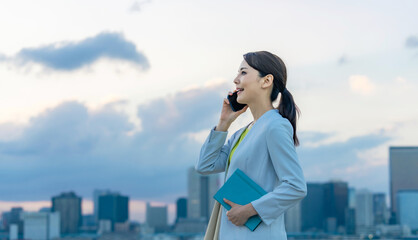 スマホ、スマートフォン、ビジネスウーマン、女性、電話、通話、ビジネス、ビジネスマン、日本人、笑顔、都市、社会、通信、5G、IoT、連絡、アプリ、サポート、問い合わせ、話す、会話、コール、屋外、ビル群、夕焼け、夜明け、夕暮れ、夕方、爽やか、働く、仕事、外出、歩く、テクノロジー、ネットワーク、電話をかける、モバイル、携帯電話、コピースペース、風景、明日、将来、展望、若い、30代、キャリアウーマン、20