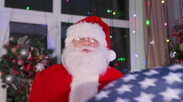 Santa dancing and waves with the US flag in a festive room decorated with a Christmas tree, garlands. Vacation in USA. Slow motion.