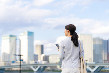 スマホ、スマートフォン、女性、ビジネスウーマン、ビジネス、日本人、キャリアウーマン、モバイル、後ろ姿、5G、都市、通信、アプリ、屋外、青空、社会、技術、テクノロジー、携帯電話、ソーシャルメディア、ソーシャルネットワーク、SNS、ビル群、都会、ユーザー、仕事、ビジネスマン、立つ、コピースペース、webサイト、インターネット、オンライン、メール、人材、20代、30代、転職、就職、展望、将来、若い、キャ