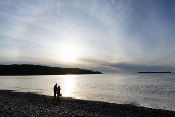 Fototapeta premium Beautiful sunrise or sunset at a beach with a corona around the sun showing an elderly retired couple with their dog in silhouette standing on the rocks looking out watching the water of the ocean