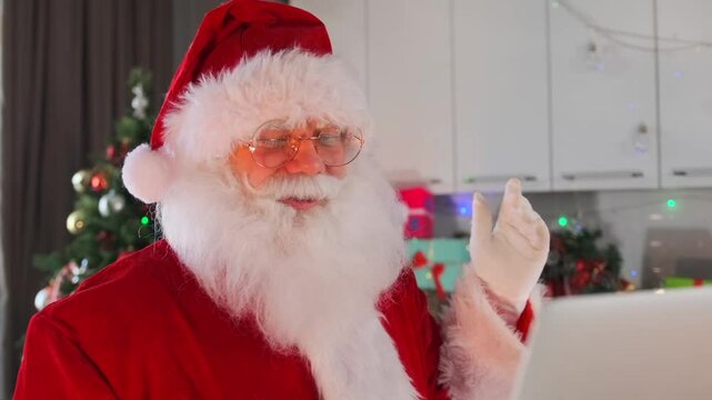 A sensible Santa Claus with a white beard chatting on a video call through a laptop standing in the middle of a room decorated with a holiday tree, garlands, gifts and a fireplace.