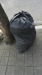 A black garbage bag that has been filled with garbage on the sidewalk during the day
