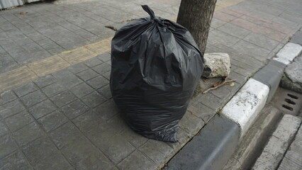 A black garbage bag that has been filled with garbage on the sidewalk during the day
