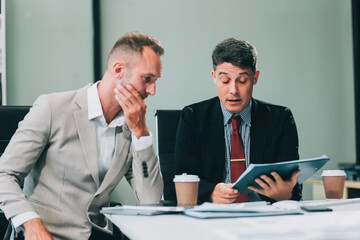 Two Unhappy tired businessman, corporate manager, sits at the workplace, tired from online work, stressed, nervous, massages the bridge of his nose, closed his eyes, experiencing a headache.