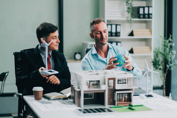 Engineers meeting about wind turbine model and house model. Renewable energy and housing development concept. Team of architects and engineers in modern office discussing design structure.