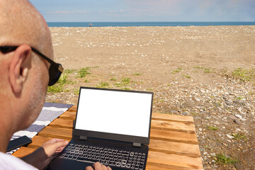 Sunny day, man in sunglasses remotely working on his laptop on a beach by the sea.digital concept