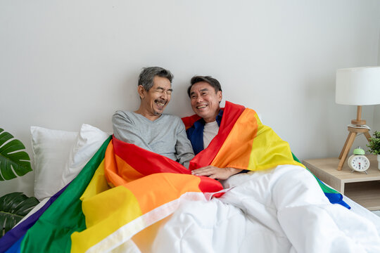 Asian elderly gay couple lying down on bed and holding rainbow flag.