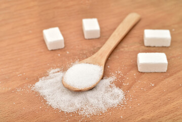 Sugar cubes and refined white sugar scattered around and in the wooden spoon on the wooden surface. Selective focus. Stop diabetes and healthy diet concept.