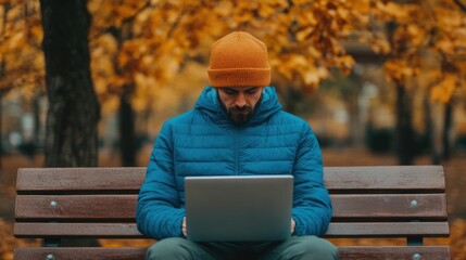 Fototapeta premium Young adult sitting on a wooden bench and using a laptop computer in an autumn city park taking advantage of the public WiFi connection and peaceful outdoor environment to work remotely