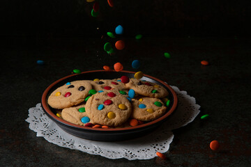 Chocolate chips falling onto a black plate with red, blue, green and yellow chocolate chip vanilla cookies