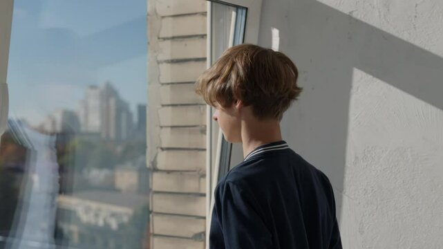 A sad abandoned boy alone at home. Sad teenage guy looking out the window, waiting for parents. Orphan teen boy waiting for adoption, feeling lonely and depressed. Stressed isolated kid feel despair 