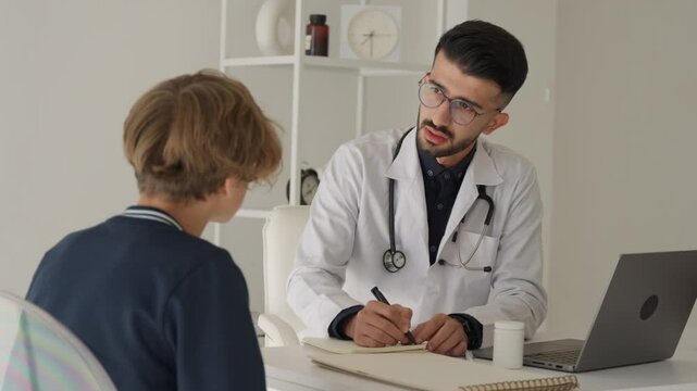 Young male doctor pediatrician talking to teen boy patient making notes in clipboard card explaining health care to school teenager fill form at medical checkup appointment