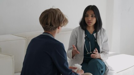 Female pediatrician listening to sick young boy patient complaints sitting in medical clinic during checkup in exam room or at home. Child doctor giving consultation in office at hospital or in house