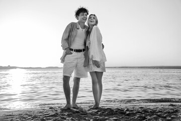 Smiling beautiful woman and her handsome boyfriend. Excited couple in casual clothes. Happy cheerful family. Female and man pose at sunrise over sea beach outdoors. Seaside in summer day, monochrome