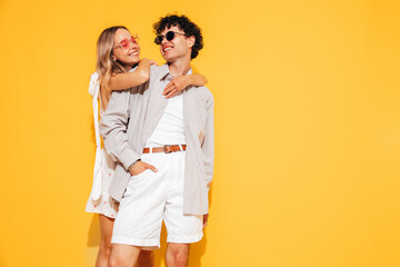 Happy beautiful romantic teenage couple. Smiling handsome caucasian man and young woman in summer clothes posing in studio, hugging sharing emotions, cheerful and happy. Isolated on yellow, sunglasses
